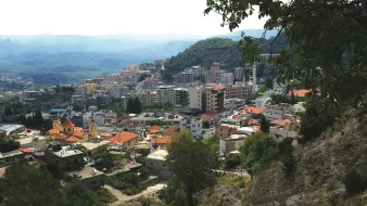 The image shows a panoramic view of a mountainous landscape with a town nestled in the valleys. The foreground features a mix of low-rise buildings, some with red rooftops, and greenery, including trees and hills. In the background, a larger urban area can be seen with taller buildings, likely indicating a more developed part of the town. The sky appears slightly overcast, with mountains extending into the distance, adding depth to the scene. The overall atmosphere suggests a blend of natural beauty and urban development.