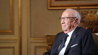 The image depicts an elderly man sitting in a formal setting. He is wearing glasses and a black suit, along with a black tie. The background features tasteful decor with intricate designs and warm colors, suggesting an elegant environment. The man appears to be engaged in thought or reflecting, with a calm and serene expression on his face.