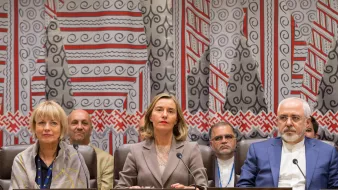 The image shows a formal setting, likely a meeting or conference related to international diplomacy. In the foreground, there are three individuals seated at a table, each identified by nameplates indicating their affiliations: the United States, the European Union, and Iran. The woman in the center, dressed in a professional outfit, appears to be the main speaker or moderator. Behind them, several other individuals are visible, suggesting a diverse group of participants in the meeting. The backdrop features a decorative pattern, adding a cultural context to the setting.