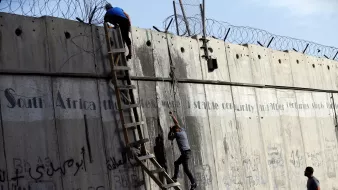 The image shows two individuals climbing a tall concrete wall, which features barbed wire at the top. A ladder is positioned against the wall, aiding their ascent. The wall bears graffiti, including text in multiple languages, and there is a phrase that mentions "South Africa" likely in the context of stability and historical struggles. The setting appears tense, capturing a moment of movement against a backdrop of urban infrastructure.