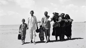 The image depicts a group of people walking across a sandy landscape. There are five adults and three children, with some carrying bundles and containers. The adults wear traditional clothing, and the children are dressed in simpler attire. The setting appears to be arid, with a clear sky and sparse vegetation in the background. The group's expressions seem serious as they move forward, suggesting a journey or migration.