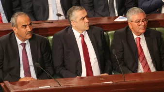 The image shows three men sitting side by side in a formal setting, likely within a legislative or governmental assembly. They are dressed in business suits, with two wearing ties—one in red and the other in a lighter color. The background suggests a gathering or meeting, with a group of people seated behind them. The scene conveys a sense of seriousness and official duty.