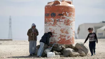 The image depicts children interacting near a large water tank. One child appears to be filling a container with water from a tap on the tank, while another child stands beside them. A third child is standing apart, looking at their phone or device. The water tank is orange and white, marked with a logo that indicates it is associated with a humanitarian organization. The surrounding area is dry and rocky, suggesting a challenging environment.