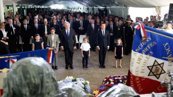 The image depicts a solemn gathering under a tent, likely at a memorial or commemorative event. In the foreground, there are several children and adults standing attentively. Prominent figures, possibly including political leaders, are present, and flags, including one with a star emblem, are displayed around them. The atmosphere appears respectful and reflective, suggesting it is a tribute to those who have passed or a celebration of their memory. Floral arrangements are visible, adding to the somber yet respectful tone of the scene.