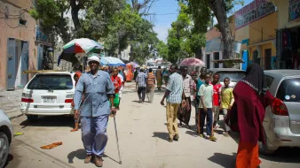 The image depicts a bustling street scene, likely in an urban area. There are several people walking along the road, some carrying umbrellas for shade. A few vehicles are parked along the sides, including cars that are mostly white. The atmosphere appears lively, with a variety of pedestrians, including a man using a walking cane. The buildings on either side are relatively simple, suggesting a community environment. Trees provide some greenery, enhancing the street's appearance. Overall, it captures a moment of daily life in a vibrant neighborhood.
