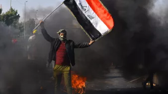 The image depicts a person holding the Iraqi flag amidst a scene of protest. The individual is wearing a mask and has their face partially covered, standing in front of billowing black smoke and flames. The background suggests a chaotic environment, likely indicative of unrest or demonstrations. The focus on the flag emphasizes national identity in the context of the protest.