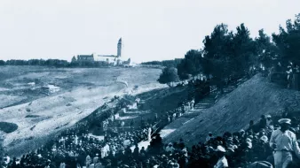 A large crowd in historical clothing gathers on a hillside, with a building in the distance.