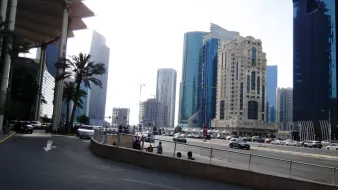 The image depicts a busy city street featuring a mix of modern skyscrapers and architectural styles. On the left, there are palm trees and a wide road leading into the city. Various cars are seen driving, indicating active traffic. The tall buildings on either side showcase glass and steel facades, reflecting light from the environment. The overall atmosphere suggests a bustling urban area with a contemporary vibe. The sky appears bright, contributing to the lively scene.