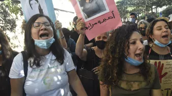 The image depicts a group of women participating in a protest. They are holding up posters, some of which feature photographs of individuals. The women appear to be chanting or shouting, expressing their emotions passionately. Many of them are wearing face masks, likely due to health precautions. The background suggests an outdoor setting with trees, indicating that the demonstration is taking place in a public space. The atmosphere seems charged with activism and determination.