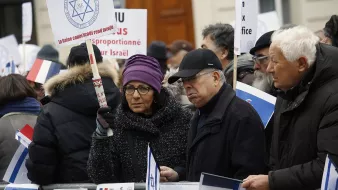The image depicts a group of protesters gathered in an outdoor setting. They are holding signs and flags, including the Israeli flag and the French flag. The attendees appear to be engaged in a demonstration, showing solidarity or expressing opinions related to Israel. Some individuals are wearing winter clothing, suggesting it is during a colder season. The signs have various messages, possibly related to political or social issues concerning Israel. The atmosphere seems serious, highlighting a significant concern among the participants.