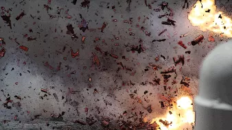 The image depicts an intense explosion, with fragments and debris scattered throughout the scene. There are bright flashes of light indicating the force of the blast, and the background appears smoky and chaotic, suggesting a highly energetic event. The overall atmosphere conveys a sense of destruction and turmoil.