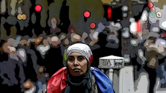 The image features a woman in the foreground wearing a white headscarf and a scarf that appears to have the colors of the French flag draped around her shoulders. She has a serious expression as she looks directly at the camera. The background is somewhat blurred and depicts a bustling crowd, suggesting a lively or busy environment, possibly a city street. The overall composition conveys a sense of solidarity or national identity amid a dynamic setting.