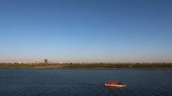 The image depicts a serene river landscape under a clear blue sky. In the foreground, a small boat with a red canopy is floating on the water, which reflects the sky. In the background, there is a sandy shore lined with greenery, and a distant cityscape can be seen, with buildings rising against the horizon. The overall scene conveys a peaceful and picturesque atmosphere.