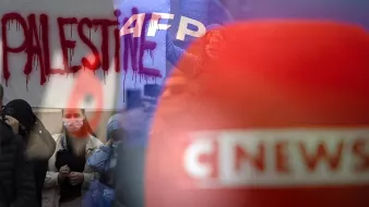 The image appears to depict a scene related to a protest or demonstration. In the background, there is graffiti that reads "PALESTINE," likely expressing support for Palestinian causes. The foreground shows blurred elements, possibly including news coverage or media logos, indicating that the event is being reported. The overall composition suggests a focus on social or political issues, drawing attention to the situation in Palestine alongside media presence.