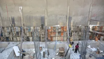The image shows a construction site with several workers engaged in various activities. There are exposed concrete walls, rebar (steel reinforcement bars) standing upright, and wooden forms for pouring concrete. The area appears to be a deep excavation where preparations for building are underway. Some materials like wood and steel are scattered around, while a few workers are busy, possibly organizing or mixing materials. The overall scene reflects typical construction activities in an urban setting.