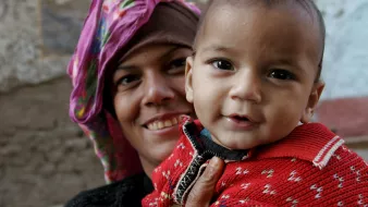 The image features a smiling woman holding a small child. The woman is wearing a colorful headscarf, and her expression is warm and cheerful. The child, who is dressed in a bright red sweater with a patterned design, looks happy and is facing the camera, showing a playful smile. The background appears to be a textured wall, suggesting an outdoor or rustic setting. The overall mood of the image is joyful and intimate, capturing a moment of connection between the woman and the child.