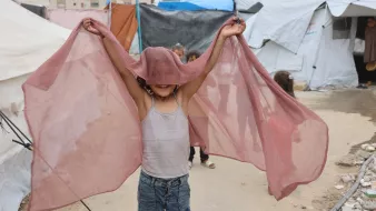 A smiling child holds up sheer fabric, playfully expressing joy in a makeshift camp.