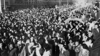 The image depicts a large crowd of people gathered in an urban setting, likely during a significant event or protest. The crowd appears to be densely packed, filling the street and surrounding areas. Many individuals are dressed in formal or semi-formal attire, indicating the seriousness of the occasion. The atmosphere seems charged, reflecting a sense of unity and purpose among the participants. Buildings line the background, and there are vehicles partially visible, suggesting a city environment. The black-and-white effect adds a historical or dramatic tone to the scene.