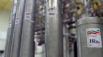 The image features tall, cylindrical metal containers labeled with the designation "IR8" and "IR8s," which suggests they are related to technological or industrial equipment, possibly in a laboratory or manufacturing setting. There are also labels indicating a flag, likely representing a country, next to the designations. The background is blurred, showing a complex arrangement of machinery or similar structures, indicating a sophisticated environment, possibly related to nuclear technology or similar fields.