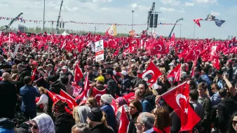 The image shows a large crowd gathered in an outdoor setting, with many people holding Turkish flags. The atmosphere appears to be lively and energetic, with individuals visibly engaged in the event. Some are holding banners, while others are raising their flags high. The background features clear skies and some structures, possibly related to the event or gathering. Overall, it depicts a significant public demonstration or celebration, likely related to a national or cultural event in Turkey.