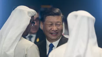 The image depicts a smiling man in a suit engaging in conversation with two individuals wearing traditional white Middle Eastern attire, including headscarves. The setting feels formal and diplomatic.