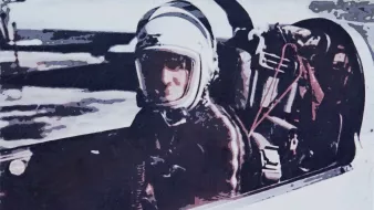 The image depicts a person seated in a cockpit, likely a pilot or astronaut. They are wearing a helmet and protective gear, suggesting they are preparing for a flight or mission. The background appears somewhat blurred, emphasizing the individual in the cockpit. The overall tone of the image conveys a sense of readiness and focus, typical of aviation or space exploration contexts.