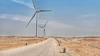 The image depicts a wide, unpaved road extending through a dry, barren landscape. On either side of the road, there are several large wind turbines standing tall against a clear blue sky. The terrain appears arid, with sparse vegetation and a sandy or rocky surface. The road is lined with small posts or markers, adding a structured element to the otherwise natural setting. The overall atmosphere conveys a sense of open space and renewable energy in a desert-like environment.