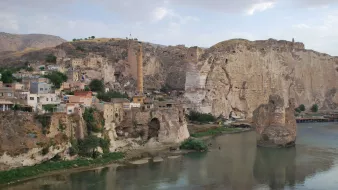 The image depicts a scenic landscape featuring a river flowing through a valley. On the left side, there are rocky cliffs and remnants of ancient structures, hinting at a historical settlement. The area appears to be a mix of natural and man-made elements, with a few buildings clinging to the hillside. In the background, there are more cliffs and a prominent tower-like structure, suggesting a rich history and possibly a fortified site. The sky is partly cloudy, adding to the serene atmosphere of the scene.