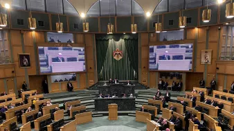 The image depicts an interior view of a legislative assembly or parliament. The chamber is spacious, featuring a semi-circular arrangement of desks for members. At the front, there is a podium where a speaker or official is addressing the assembly. Large screens on either side display information or the speaker to the attendees. The walls are adorned with drapes and emblems, contributing to a formal atmosphere. Members of the assembly are seated, engaged in discussion or listening attentively. The overall setting is one of a governmental deliberation space, likely during a session or meeting.