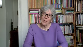The image features an elderly woman with gray hair and glasses, sitting at a table. She is wearing a purple sweater and appears to be engaged in conversation or sharing her thoughts. In the background, there are bookshelves filled with various books, creating a colorful and organized backdrop. The setting suggests a cozy and intellectual atmosphere.