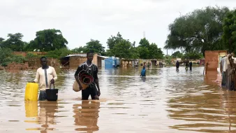 The image depicts a flooded area, where individuals are walking through standing water. Two men are central in the foreground; one is carrying a bucket, while the other holds a rolled-up mat. In the background, makeshift shelters or homes are partially submerged, surrounded by trees and greenery. The scene reflects the impact of flooding, with water covering the ground and affecting the surrounding environment. The sky is overcast, suggesting ongoing adverse weather conditions.