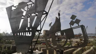 The image depicts two large, metallic sculptures or structures under construction or installation in an industrial outdoor setting. The sculptures show abstract forms, one resembling a stylized letter or shape and the other possibly depicting a ship or vessel. The background features a harbor with cranes and construction materials scattered around, suggesting an active worksite. The sky is partly cloudy, adding to the overall atmosphere of a busy environment.