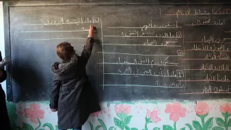 The image depicts a classroom scene where a student is writing on a blackboard. The blackboard has lines of text in Arabic, while the bottom of the board is decorated with colorful flower drawings. The student, wearing a coat, is focused on writing, and there are other individuals in the background, potentially teachers or classmates. The setting appears to be an educational environment.