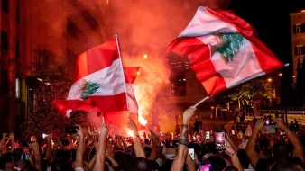 The image depicts a large gathering of people celebrating, likely in an urban setting. Many individuals are holding flags of Lebanon, which feature a red and white design with a green cedar tree. The atmosphere appears vibrant and energetic, with colorful smoke and lights adding to the scene. People are also raising their phones to capture the moment, suggesting a significant event or celebration taking place.