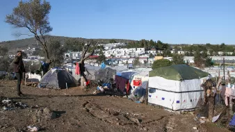The image depicts a makeshift settlement in a rural or semi-urban area. Numerous tents are arranged on a slope, suggesting a temporary living situation. The landscape features sparse vegetation and a few trees, while in the background, there are more established buildings, possibly residences or facilities. The scene conveys a sense of hardship, with signs of disarray and poverty surrounding the tents. The sky is clear with little cloud cover, indicating a sunny day. A person stands in the foreground, observing the surroundings.
