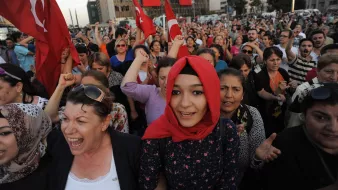The image depicts a large crowd gathered, likely in an outdoor setting, with many individuals holding Turkish flags. People appear to be passionately engaged, with expressions of enthusiasm and solidarity. Some individuals are wearing traditional attire, while others wear modern clothing. The background features a building adorned with large banners, adding to the sense of national pride and collective emotion in the scene. The atmosphere seems charged with energy, suggesting a demonstration or celebration of national significance.