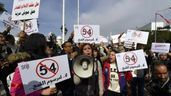 The image depicts a protest scene with a group of people holding up signs and banners. Some demonstrators are prominently featuring a sign with the number "54" crossed out, suggesting opposition to a specific law or regulation. One woman is speaking into a megaphone, leading the crowd in their protest. The atmosphere appears energetic and determined, with a focus on raising awareness or voicing their concerns regarding a particular issue.