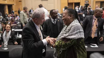 The image depicts a moment of interaction between two individuals at a conference or event. Both people are smiling and engaged in a handshake, suggesting a friendly and positive exchange. The person on the left is wearing a suit, while the individual on the right is dressed in traditional or cultural attire with a colorful shawl. In the background, there are other attendees, some seated and others standing, indicating a busy atmosphere typical of a conference. The setting appears to be indoors, likely within a large hall or meeting room.