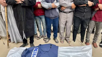 A blue "PRESS" jacket rests on a white sheet, surrounded by people with arms crossed.