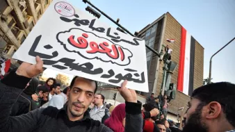 The image depicts a crowd gathered in an urban setting, likely during a protest or demonstration. In the foreground, a man holds a large sign with Arabic text, which translates to "Our demands are not to be ignored." In the background, there is a building displaying the Egyptian flag, suggesting a context related to political or social issues in Egypt. The scene conveys a sense of activism and public expression, with people rallying for their rights or changes in governance.