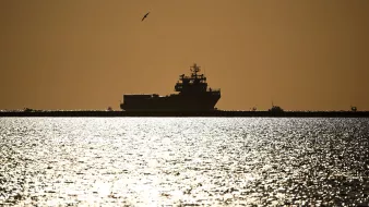 The image depicts a silhouette of a large ship on water during sunset or sunrise. The scene is bathed in a warm golden light, creating a dramatic contrast between the dark shape of the ship and the shimmering, reflective surface of the water. A seagull is seen flying in the sky, adding to the serene atmosphere of the moment. The horizon appears distant, enhancing the feeling of calmness and stillness in the scene.