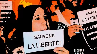 The image depicts a group of people holding up signs with messages in French. The main focus is on two individuals in the foreground. One is wearing a hijab and both are raising their signs high, which read "Sauvons la liberté!" (Save Liberty!). The atmosphere suggests a protest or rally, with a sense of solidarity and determination among the participants. The background is filled with more people and signs, contributing to the overall theme of advocating for freedom.