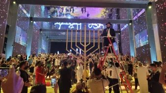 The image depicts a lively event taking place in a large, well-lit venue. In the center, there is a large menorah, which is a symbol associated with Hanukkah. A person is standing on a ladder, seemingly preparing to light the menorah. Surrounding the menorah, a diverse crowd is gathered, with some individuals clapping or taking photos. The background features colorful lights and projections, enhancing the festive atmosphere of the gathering. The overall scene reflects a sense of celebration and community.