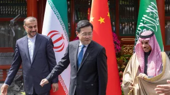 The image features three men standing together, with national flags of Iran, China, and Saudi Arabia in the background. The person on the left is wearing a suit with an Iranian flag pin, the central figure is dressed in a blue suit, and the individual on the right is wearing traditional Saudi attire. They appear to be engaged in a conversation or moment of interaction, suggesting a diplomatic or friendly meeting. The setting seems to be outdoors, possibly at a state event or formal gathering.