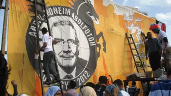 The image shows a group of people working together to paint or prepare a large mural or banner. The mural features a prominent circular design with the name "Abdel-Moneim Abol Fotoh" and the year "2012." There is a dragon motif in the background, which suggests a strong and bold theme. Some individuals are on ladders, actively engaged in applying paint, while others are gathered below, possibly helping or observing. The people are dressed in a variety of clothing styles, and there are some individuals wearing colorful wigs, indicating a festive or celebratory atmosphere. The backdrop is bright orange with various designs.