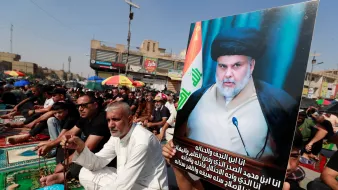 The image depicts a large gathering of people, seated on mats in an outdoor setting, likely in a city square. Many attendees are dressed in black, which is often associated with mourning or commemorative events. One man stands out, holding a prominent poster featuring a portrait of an individual wearing traditional attire, along with a flag in the background. The atmosphere suggests a significant religious or political event, characterized by unity and solemnity among the participants.