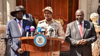 The image shows a group of three men standing behind a wooden podium. The man on the left is wearing a black hat and a suit with a red tie. The man in the center is wearing a military uniform with various insignia and a cap. The man on the right is dressed in a dark suit. They appear to be at an official event, likely addressing the media, with microphones positioned in front of them and a decorative building in the background.