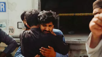 Two men embrace tightly, one looks visibly emotional, surrounded by others in a somber setting.