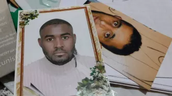 The image shows two photographs. One is a formal portrait of a man with short hair, wearing a light-colored sweater. The background contains decorative elements like flowers. The second photo appears to be a more candid or casual shot of another man, who has dark, curly hair and a neutral expression. Both photos are placed together on a surface that includes some papers and miscellaneous items.