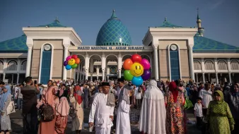 The image depicts a large crowd gathered outside the National Mosque of Al-Akbar in Surabaya, Indonesia. The mosque features a prominent architectural style with a blue and green patterned dome. In the foreground, people of various ages and attire are seen, many wearing traditional Islamic clothing. Some individuals are holding colorful balloons with smiling faces, adding a festive atmosphere to the scene. The sky is clear and blue, suggesting a bright and sunny day.